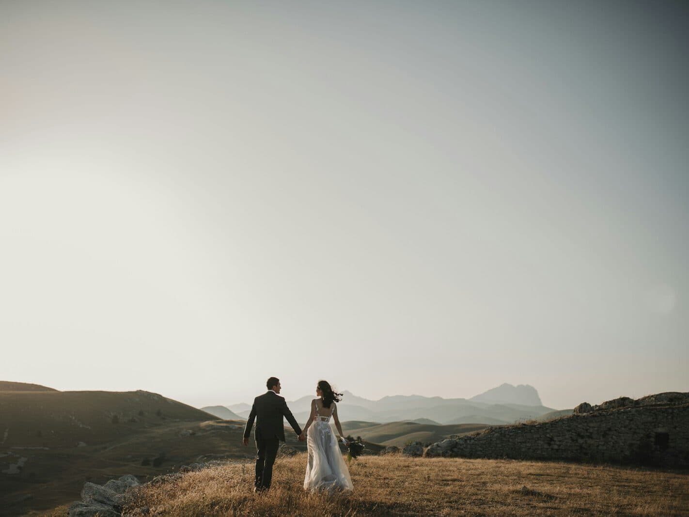 Una pareja recién casada camina de la mano por un prado de montaña al atardecer.